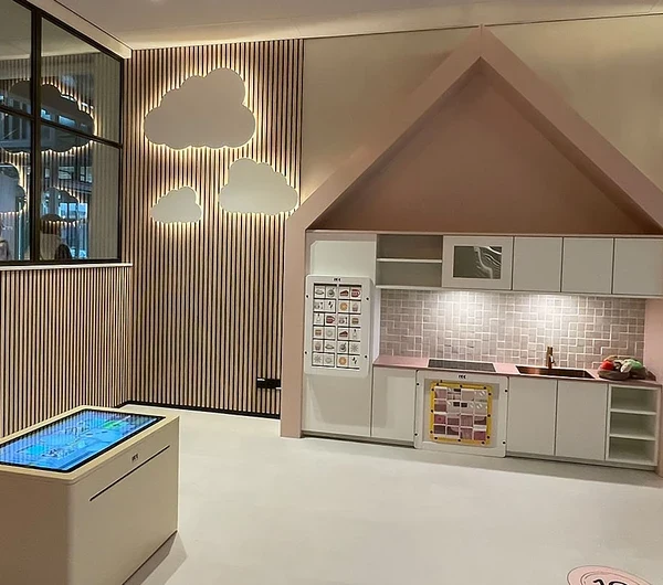 Een sfeervol ingerichte kinderhoek met een speelkeuken in een huisvormig thema, interactieve vloer met hinkelcirkels en een digitale speeltafel. De wand is versierd met houten latten en wolkvormige lichtpanelen. De ruimte is warm verlicht en bevat speelse elementen zoals een houten boomstructuur aan het plafond en retro lampen.