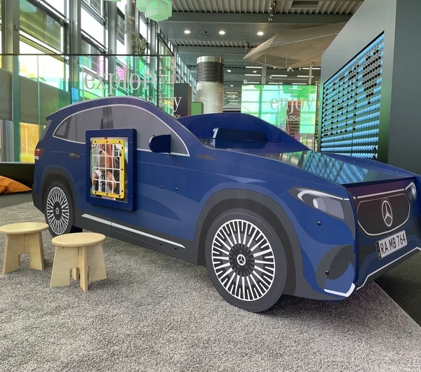Blauw Mercedes-auto speelmeubel met wandspel, houten krukjes en kleurrijke zitzakken op de achtergrond, geplaatst in de kinderhoek van het Mercedes-Benz Kundencenter in een moderne showroomomgeving.