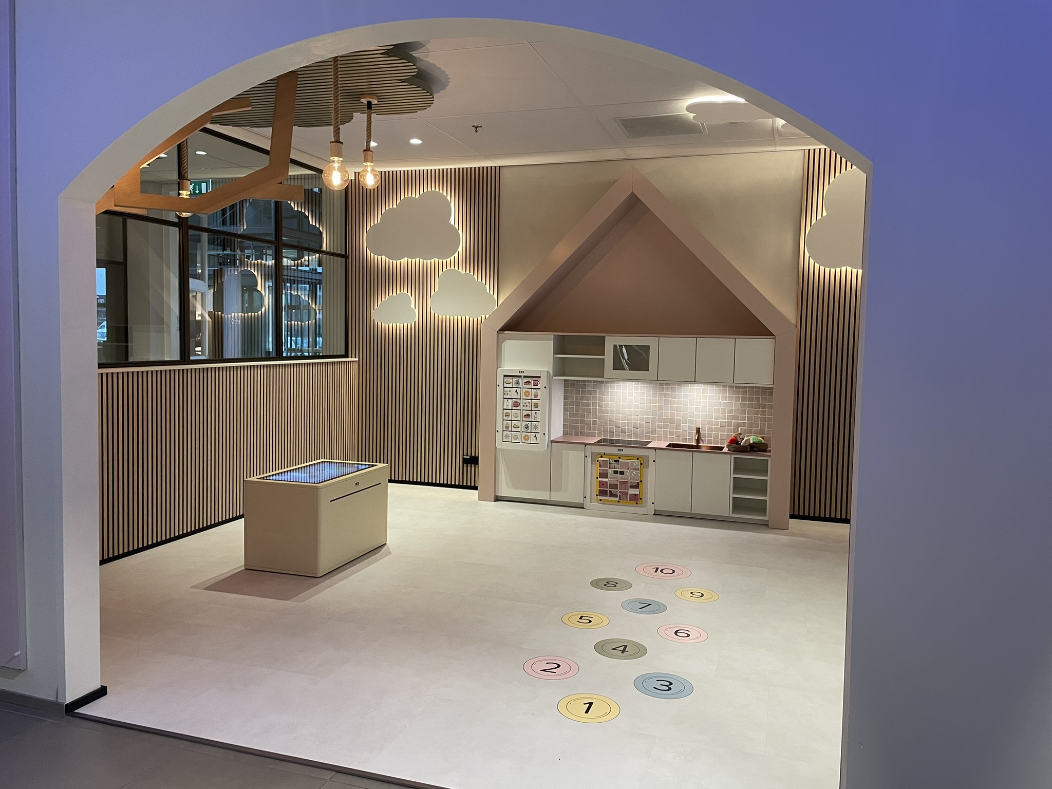 Overzichtsfoto van de kinderhoek bij Tieleman Keukens, ingericht door IKC. De ruimte bevat een speelkeuken in een huisvormige nis, een interactieve speeltafel, wanddecoratie in de vorm van wolken en kleurrijke cijfercirkels op de vloer voor speelse activiteiten. De ruimte is zichtbaar vanuit een boogdoorgang naar de showroom.