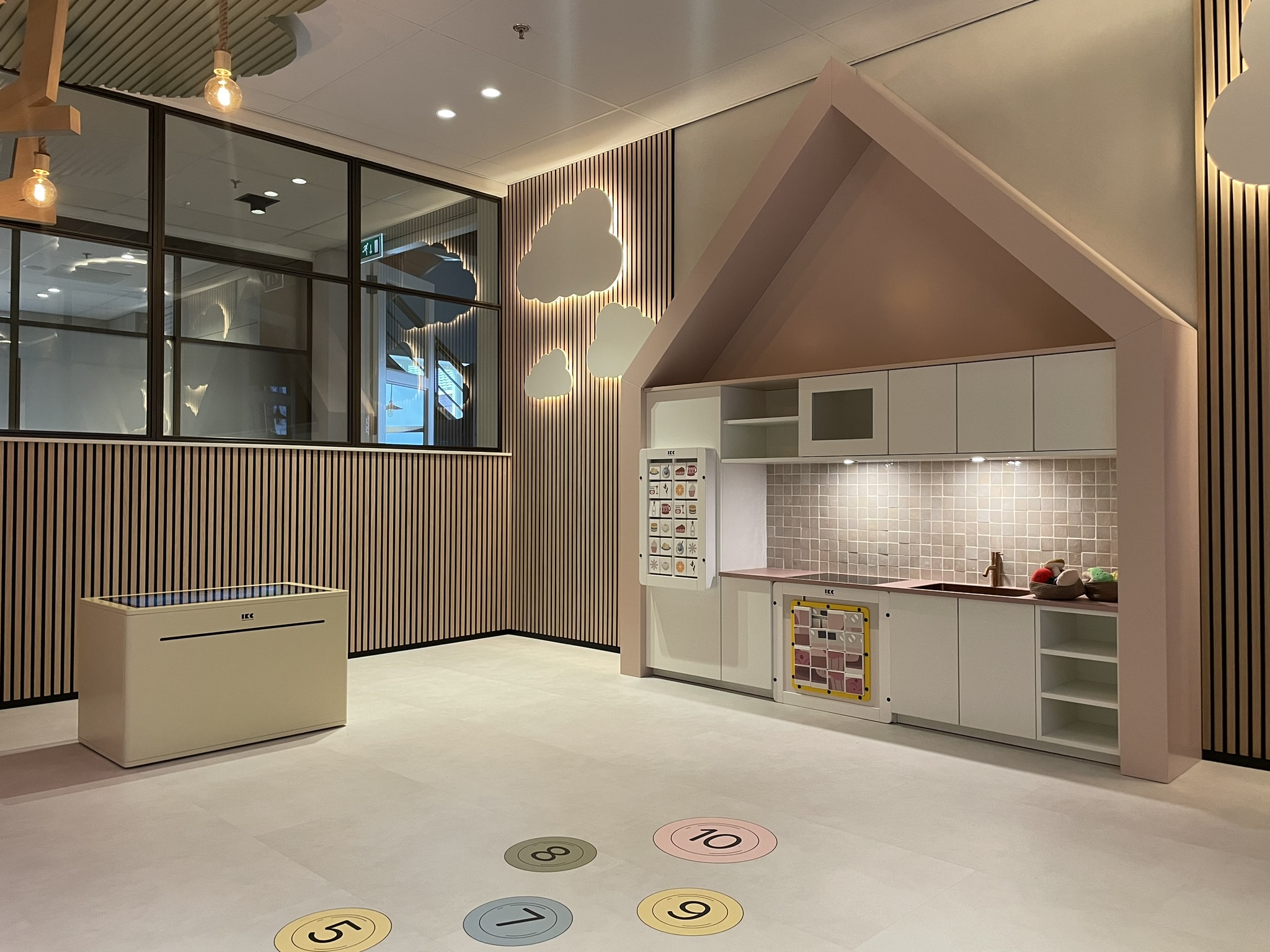 Kinderhoek in de showroom van Tieleman Keukens, ingericht door IKC, met een kindvriendelijke speelkeuken in huisvorm, interactieve speeltafel en speelcirkels op de vloer. De ruimte heeft een warme uitstraling met houten wandpanelen en verlichte wolkdecoratie.