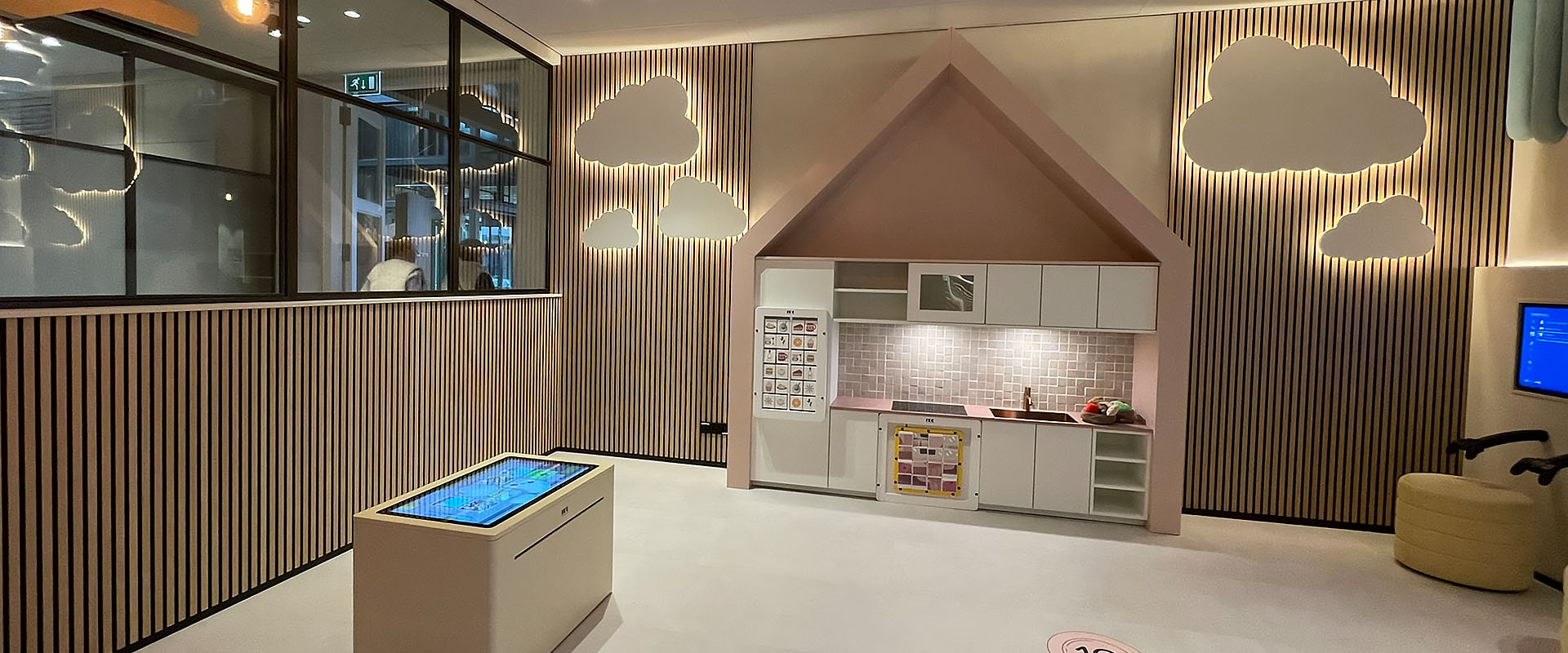 Speelse kinderhoek met interactieve elementen in een modern interieur Een sfeervol ingerichte kinderhoek met een speelkeuken in een huisvormig thema, interactieve vloer met hinkelcirkels en een digitale speeltafel. De wand is versierd met houten latten en wolkvormige lichtpanelen. De ruimte is warm verlicht en bevat speelse elementen zoals een houten boomstructuur aan het plafond en retro lampen.