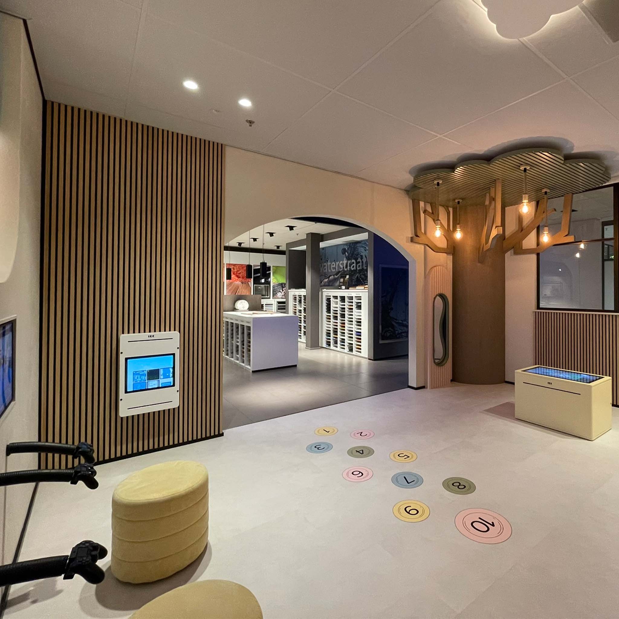 Speel en ontdek in de kinderhoek bij Tielemans Keukens Kinderhoek bij Tielemans Keukens met interactieve schermen, zachte zitpoefs, digitale speeltafel en speelse decoratie zoals een boomstructuur met hanglampen. De ruimte grenst aan de showroom en is ontworpen als belevingsplek voor kinderen terwijl ouders keukens bekijken.