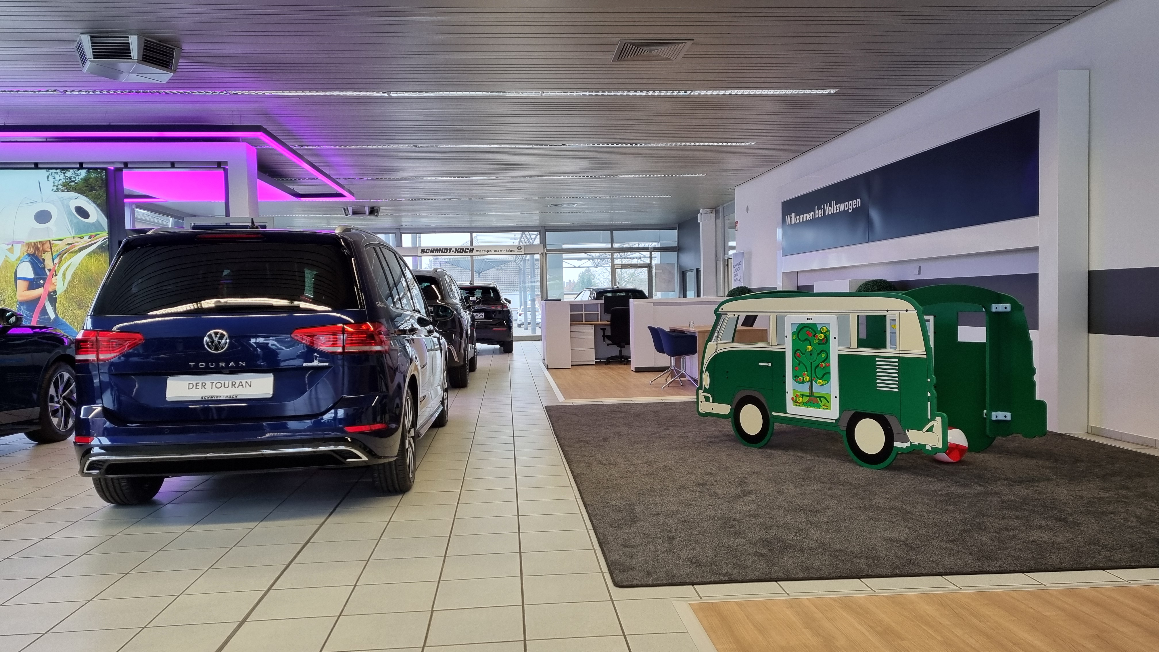Volkswagen showroom met meerdere auto's, waaronder een Touran, en rechts een speelhoek in de vorm van een groene IKC VW Van. De speelhoek is geplaatst op tapijt en sluit visueel aan op de omgeving.