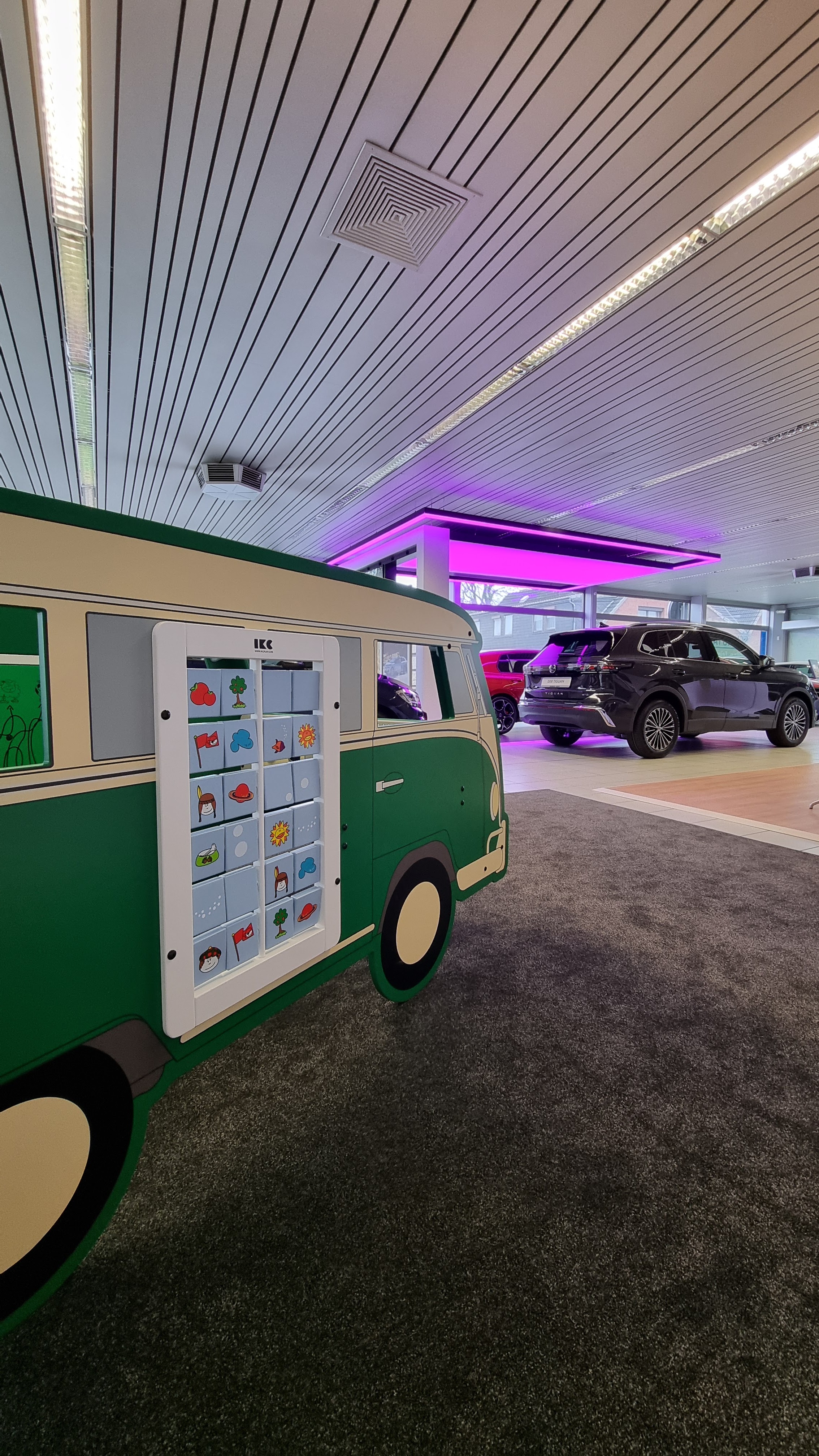 Groene IKC speelbus met flippaneelwand in Volkswagenstijl, geplaatst op tapijt in een moderne showroom met diverse auto’s op de achtergrond en opvallende paarse verlichting.