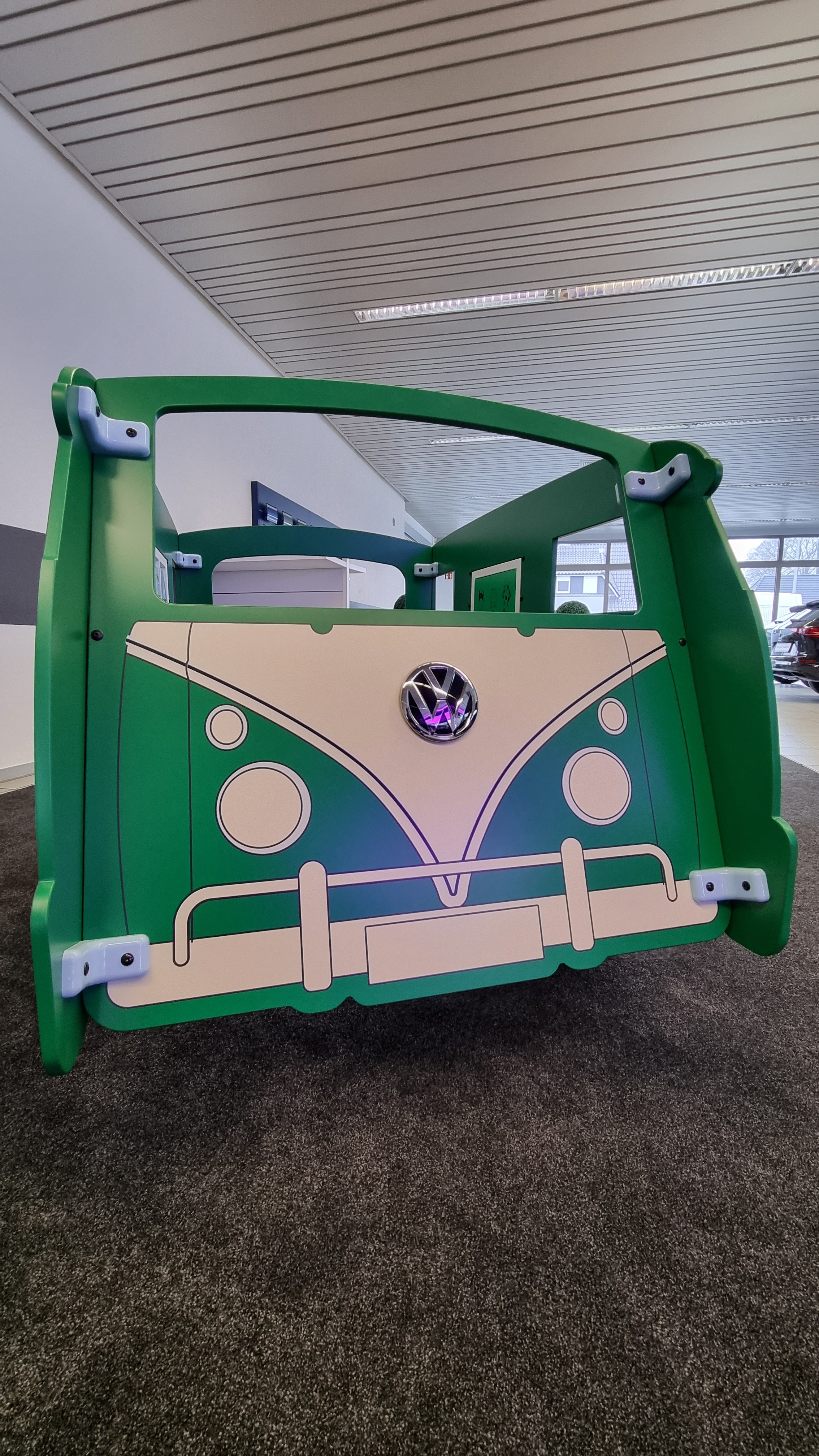 De voorkant van een groene IKC speelbus in Volkswagenstijl, geplaatst in een klantgesprekzone in de showroom van Autohaus Schmidt + Koch. Op de achtergrond een welkomstbord met de tekst "Willkommen bei Volkswagen".
