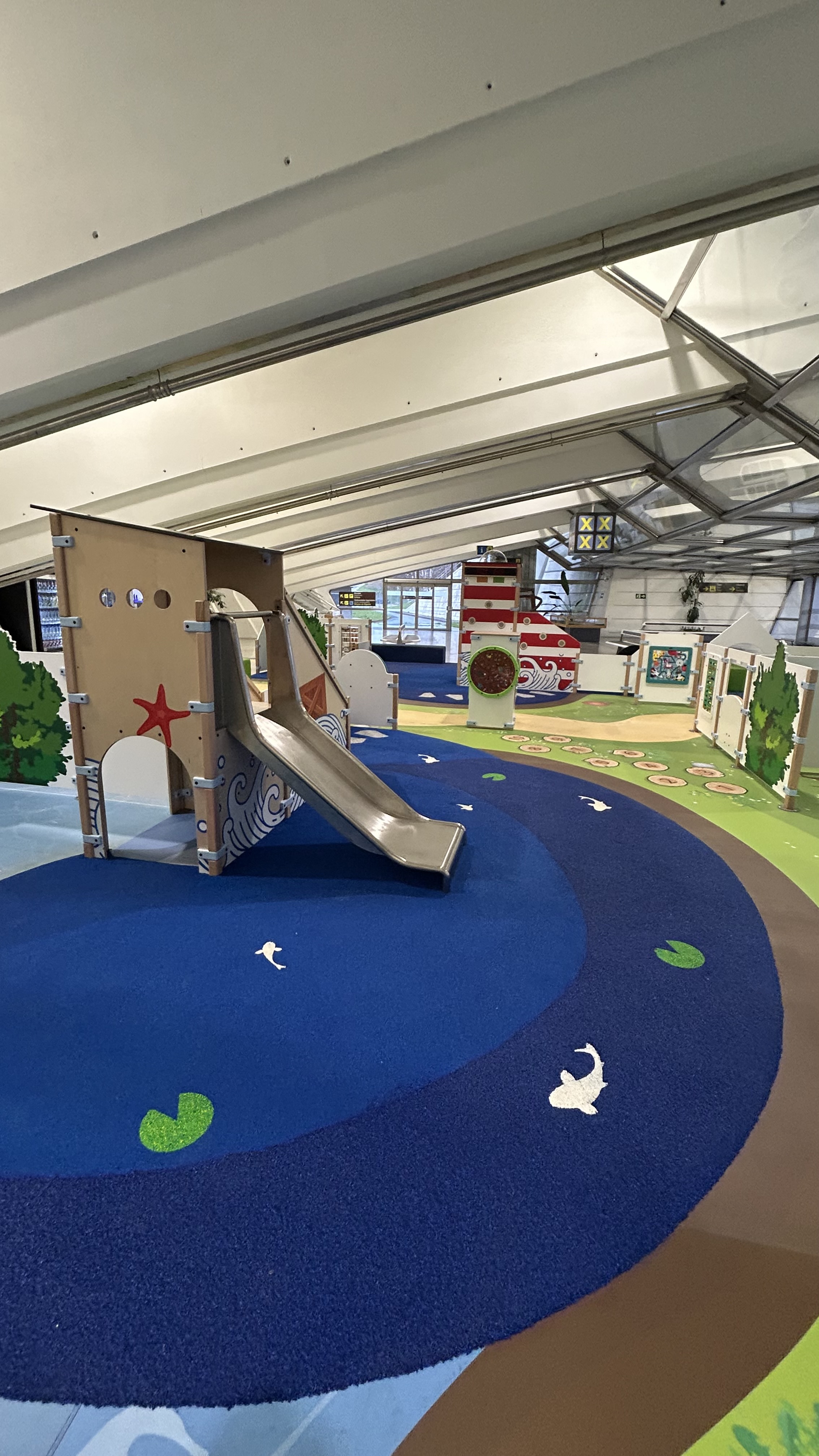 Kinderspeelhoek op Bilbao Airport met blauwe speelkussenvloer, glijbaan in maritiem thema, vuurtoren-speelelement en diverse interactieve spellen in een lichte luchthavenhal.