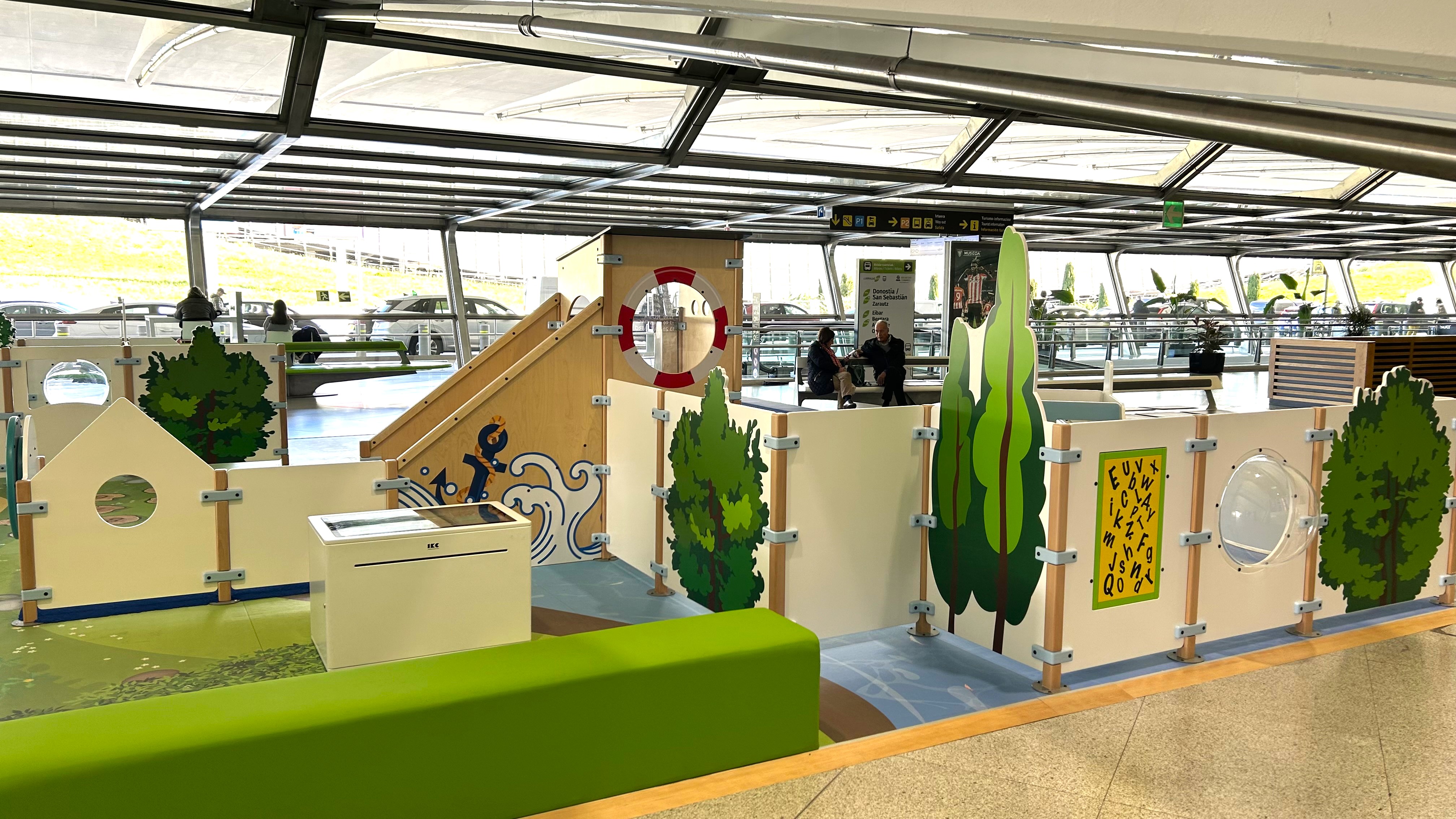Veilig en speels wachten in Bilbao Airport – kinderspeelhoek in maritieme stijl "Kindvriendelijke speelhoek op Bilbao Airport met maritiem thema, inclusief klimelementen, interactieve spellen en zitbank voor ouders in een lichte vertrekhal."