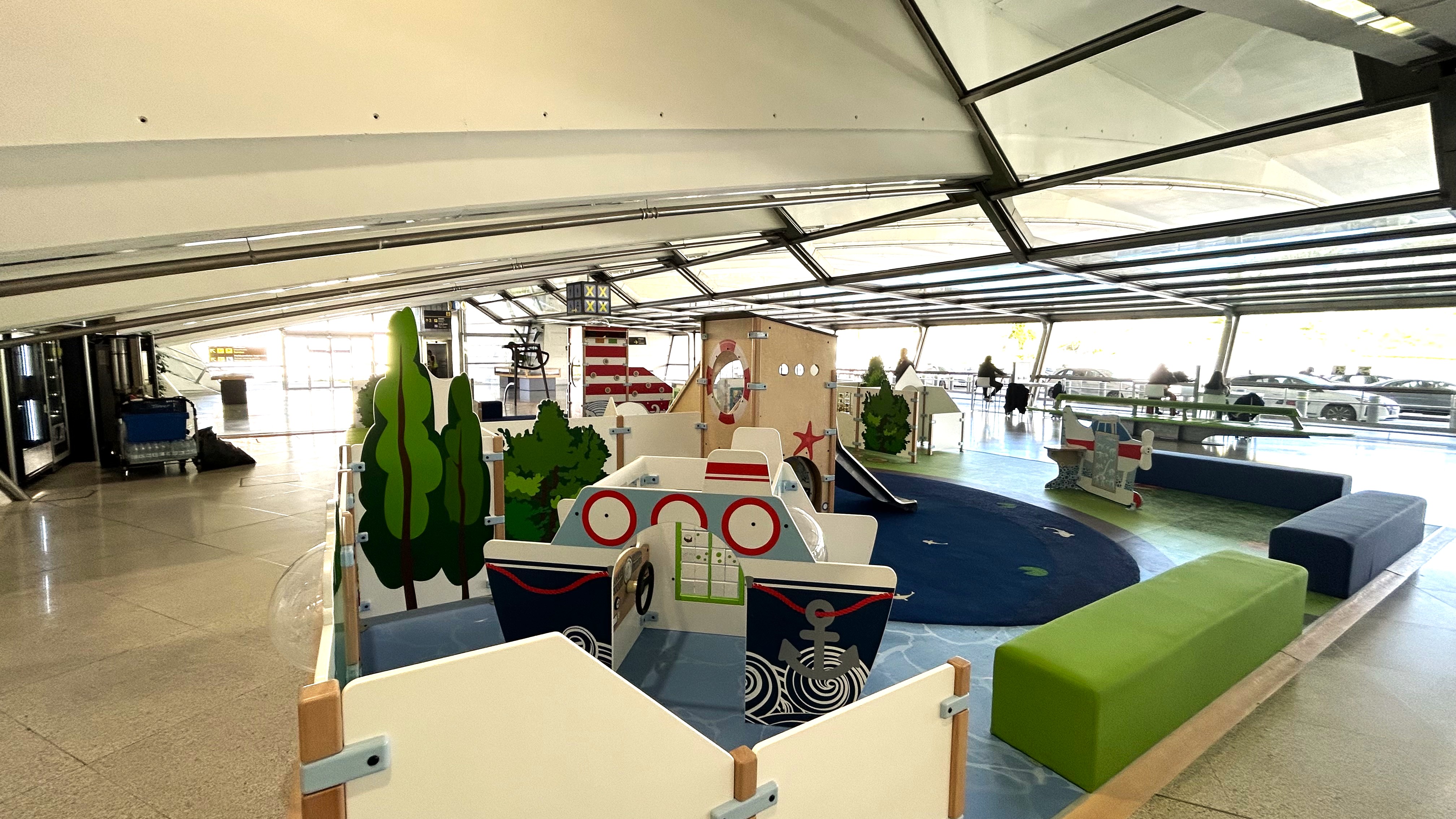 Speelzone op Bilbao Airport met maritiem thema, inclusief speelschip, vuurtoren, glijbaan en vliegtuigje, omgeven door comfortabele zitbanken voor ouders in een lichte vertrekhal.
