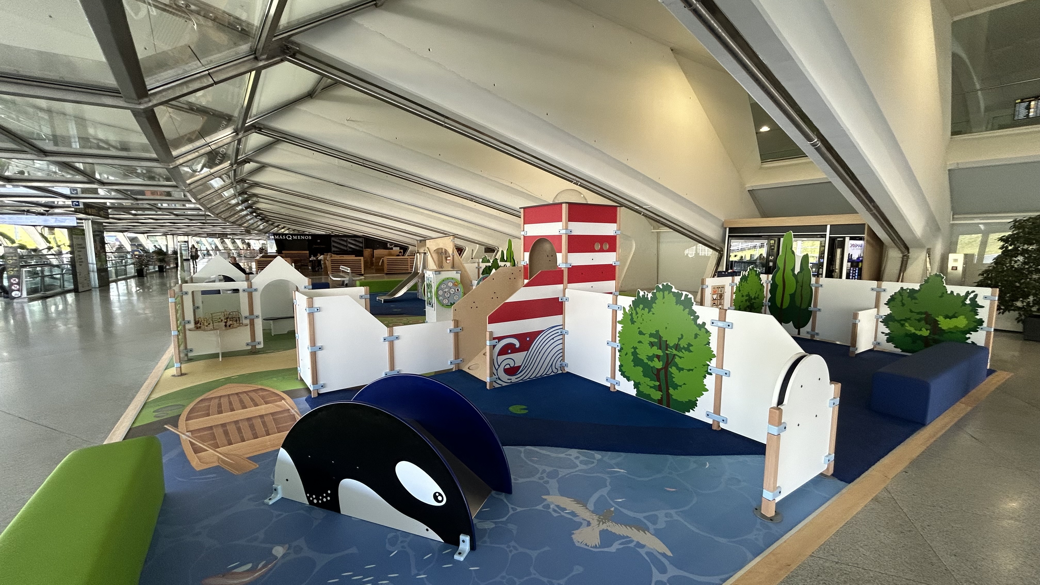 "Overzicht van de kinderspeelhoek op Bilbao Airport, met maritiem thema zoals een vuurtoren, houten roeiboot en walvis-speelelementen, ontworpen voor spel en ontspanning tijdens het wachten."