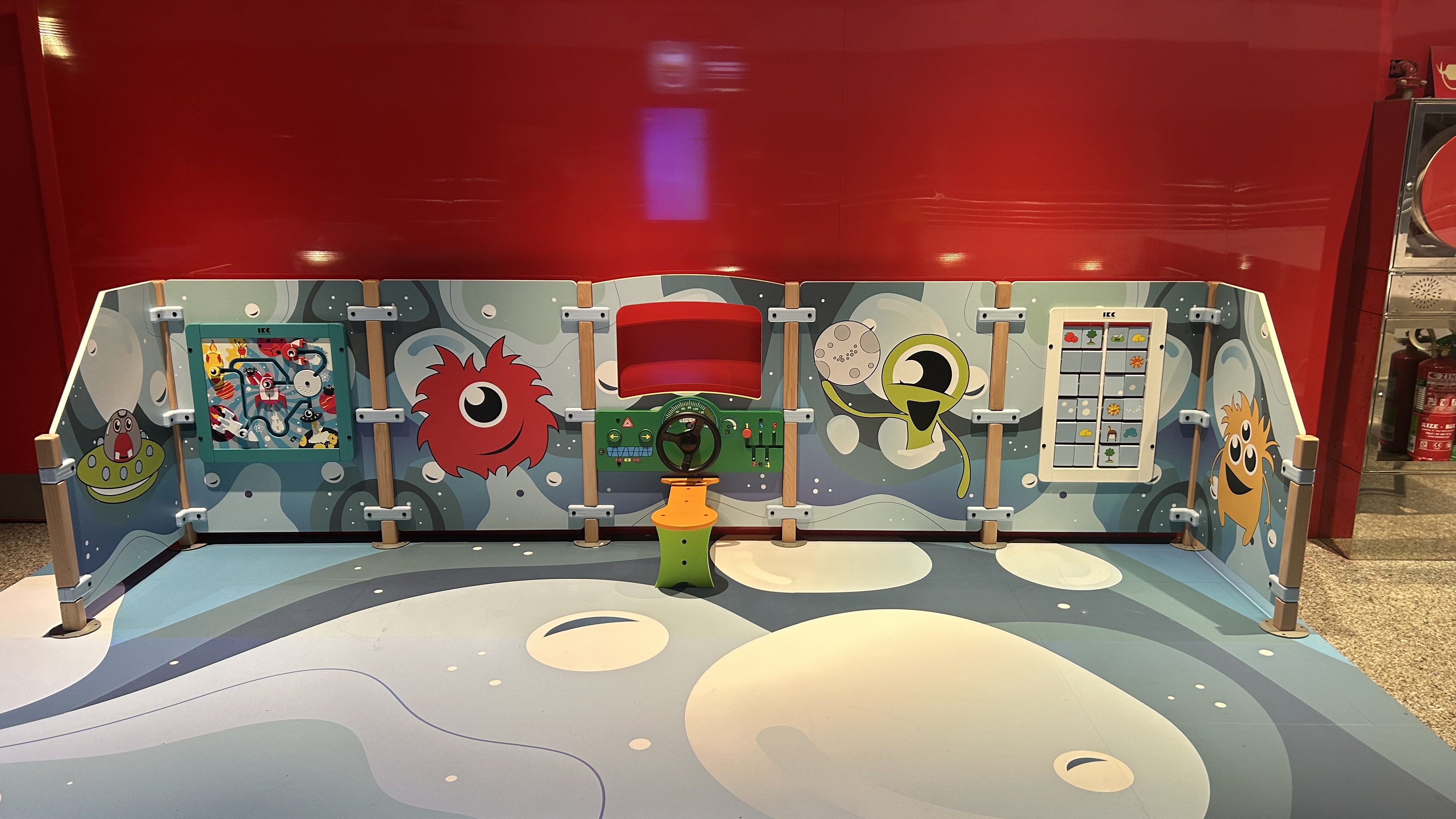 Kinderspeelwand met ruimte-thema, kleurrijke buitenaardse figuren en interactieve spelelementen zoals stuurwiel, schuifpuzzels en spelpanelen, geplaatst tegen een rode wand.