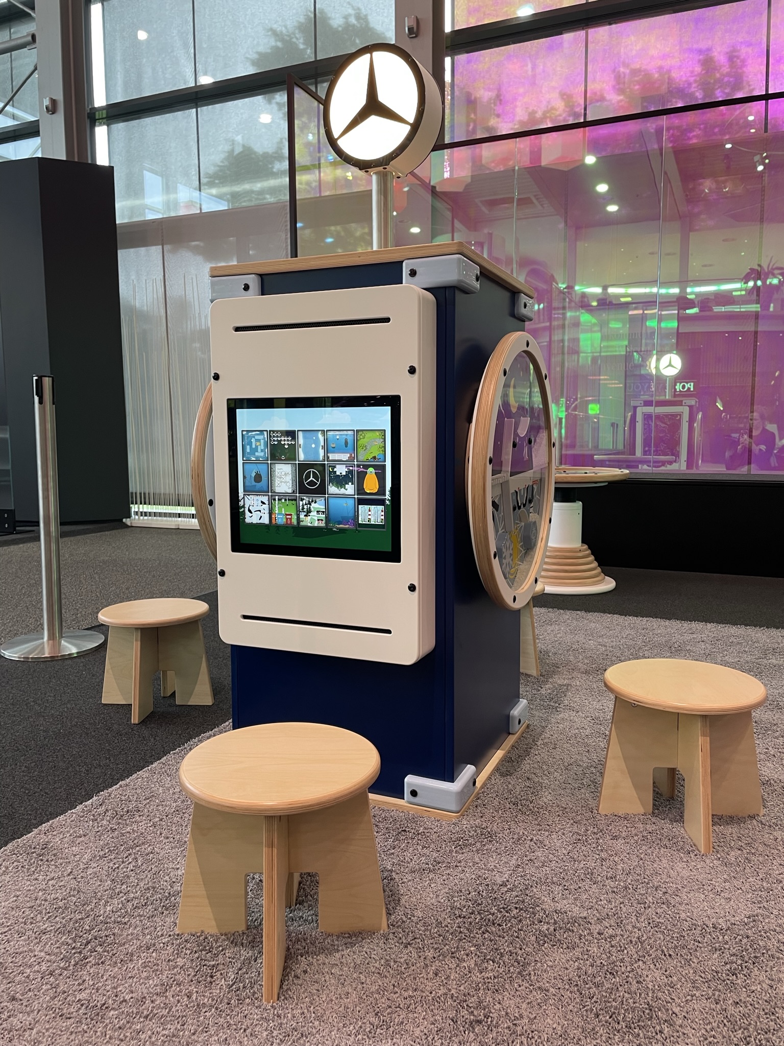 Interactief speelmeubel in Mercedes-Benz Kundencenter met digitaal touchscreen vol educatieve spelletjes, houten krukjes en het Mercedes-logo bovenop, geplaatst in een kleurrijke en moderne showroomomgeving.