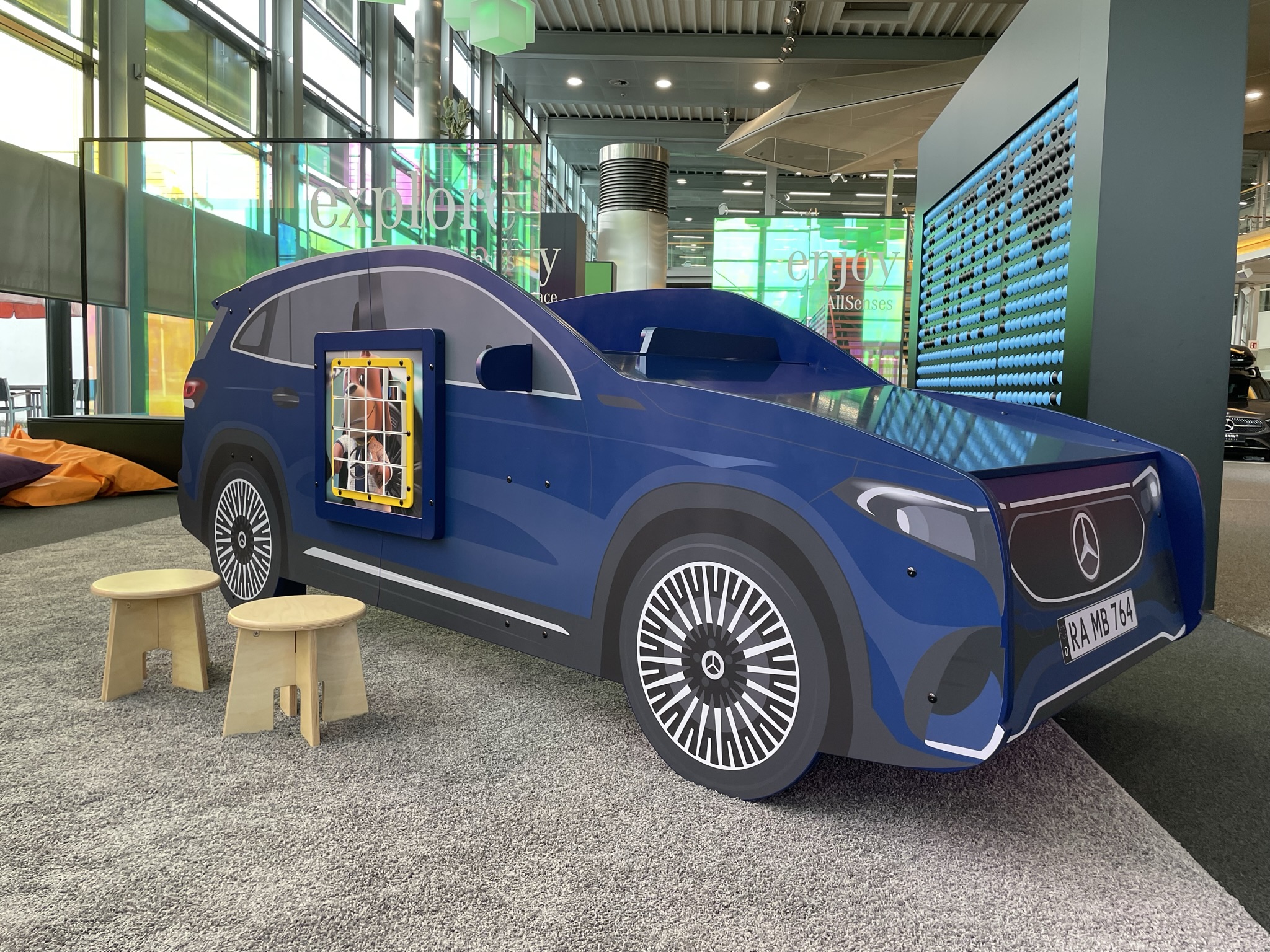 Blauw Mercedes-auto speelmeubel met wandspel, houten krukjes en kleurrijke zitzakken op de achtergrond, geplaatst in de kinderhoek van het Mercedes-Benz Kundencenter in een moderne showroomomgeving.