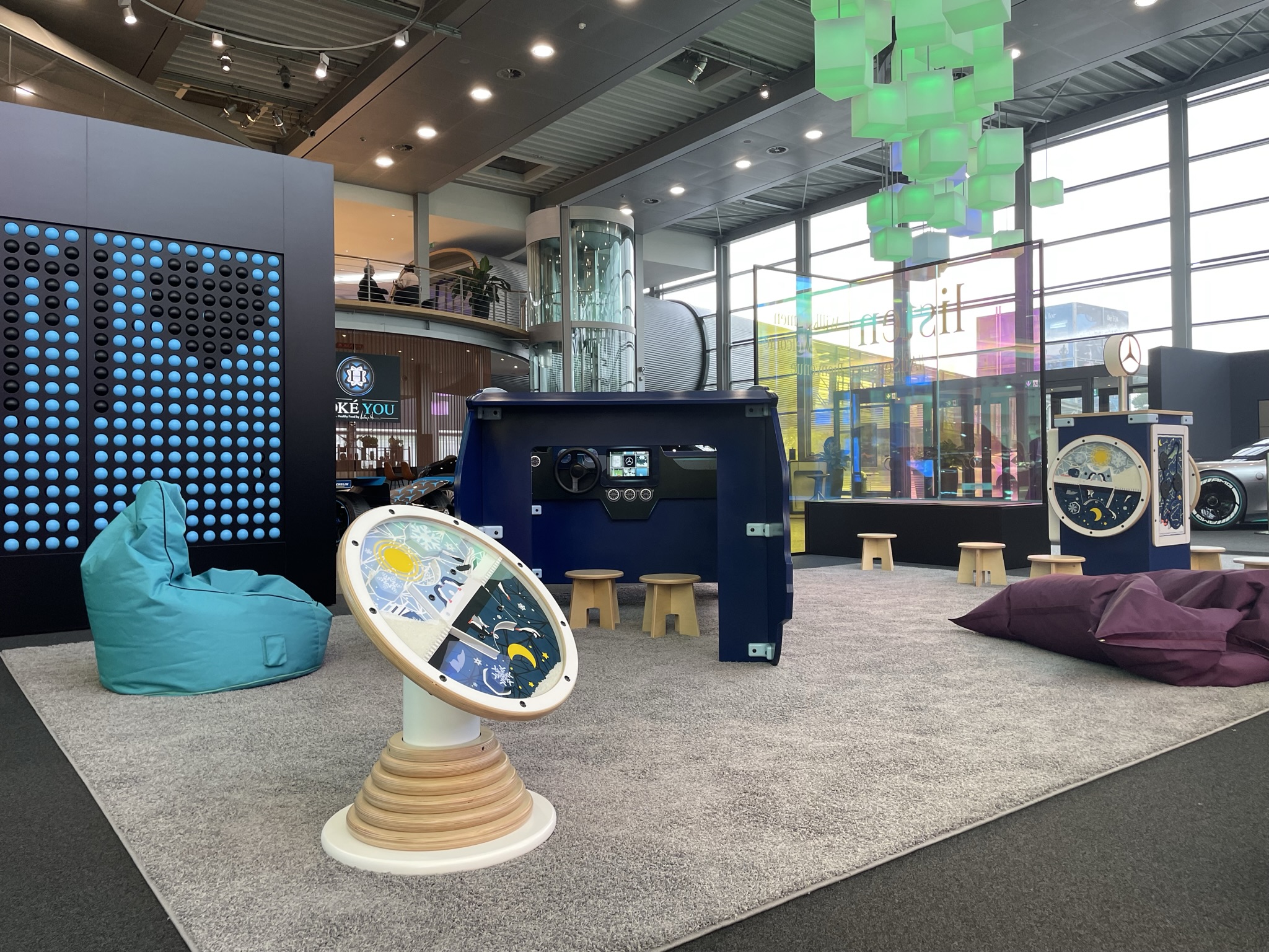 Kinderhoek in Mercedes-Benz Kundencenter met diverse speelobjecten, waaronder een blauw dashboard-speelmeubel, interactieve wandspellen, grote turquoise en paarse zitzakken en kleurrijke verlichting in een moderne showroomomgeving.