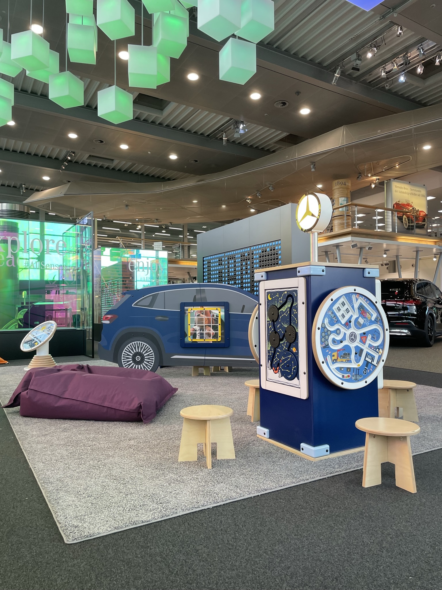 Kinderhoek in Mercedes-Benz Kundencenter met blauw auto-speelmeubel, interactief speelstation met Mercedes-logo, diverse wandspellen, houten krukjes en paarse zitzak, in een kleurrijke en moderne showroomomgeving.