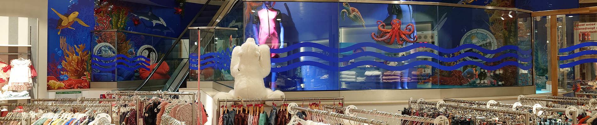 Magische onderwater-speelwereld bij Modehaus Baltz Kinderafdeling van Modehaus Baltz met kleurrijk onderwaterthema, inclusief muurschilderingen van een octopus, schildpad en vissen, naast de roltrap en kledingrekken