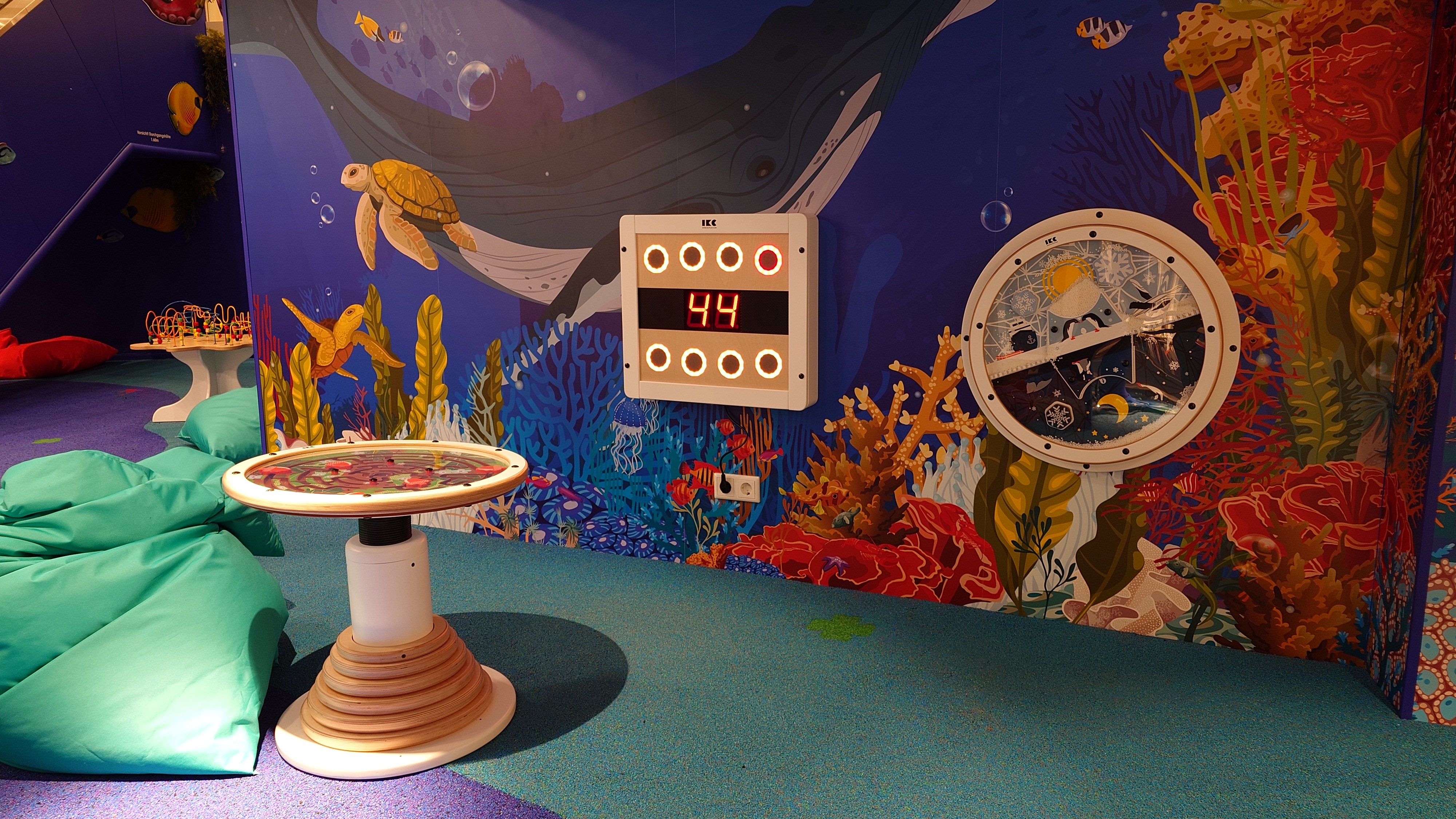 Kinderhoek in oceaanthema met digitale wandspellen, speeltafel, kleurrijke onderwaterillustraties en zachte zitzakken, ontworpen door IKC in een winkelcentrum.