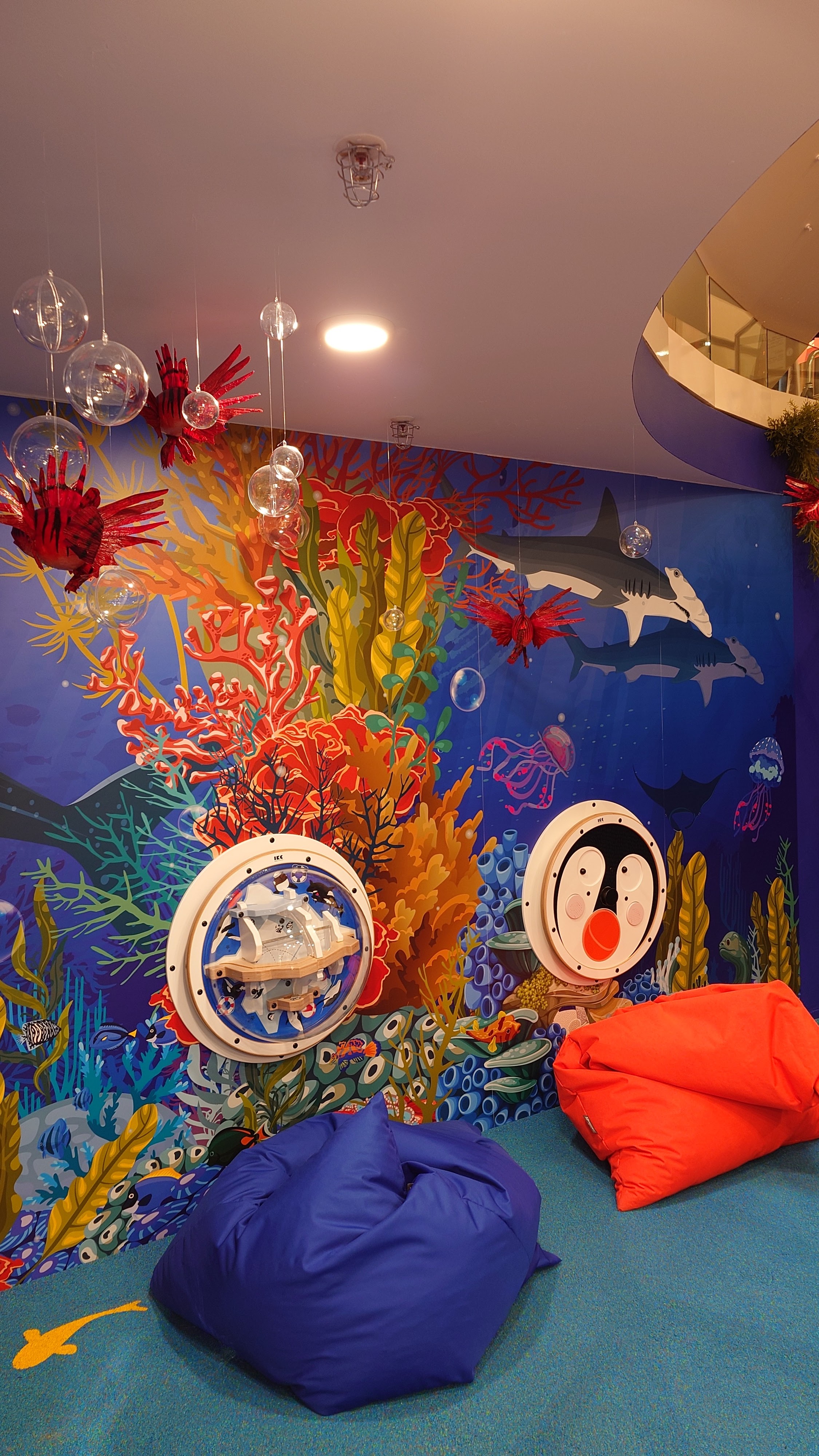 Kinderhoek in oceaanthema met interactieve wandspellen, kleurrijke koraal-illustraties, zachte zitzakken en speelse details, ontworpen en gerealiseerd door IKC in een winkelomgeving.