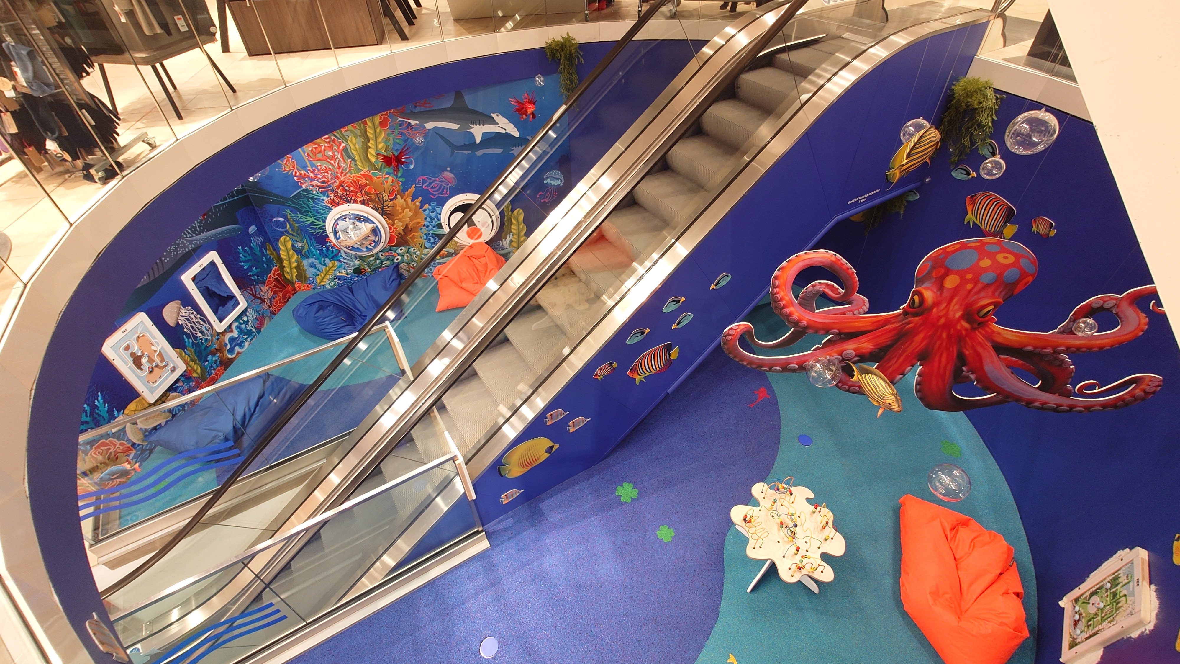 Kinderhoek in oceaanthema rondom een roltrap, met wandspellen, kleurrijke onderwaterillustraties, grote octopusdecoratie en zachte zitzakken, ontworpen door IKC in een winkelcentrum.